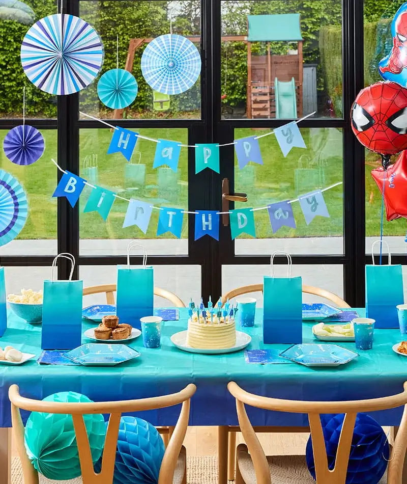 Blue birthday party table with a birthday cake, gift bags, themed decorations, hanging paper fans, and superhero balloons.